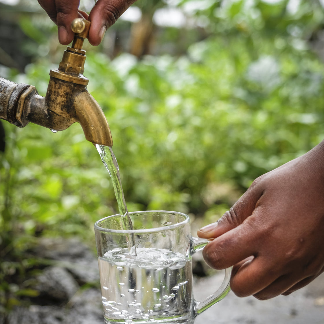 Person filling a glass with water from a brass faucet outdoors.